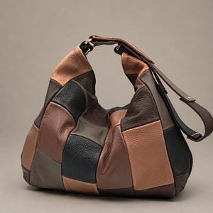 Puede incluir: Un bolso de estilo hobo confeccionado con un mosaico de paneles de cuero en varios tonos de negro, marrón oscuro, tostado y marrón claro. El bolso cuenta con una sola correa integrada para el hombro y una silueta holgada y redondeada.