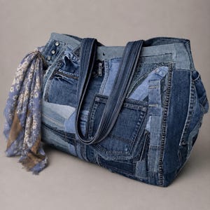 Può includere: Una grande borsa tote realizzata con vari pezzi di tessuto denim in diverse tonalità di blu, dotata di molteplici tasche visibili, cuciture e due robuste tracolle. Una sciarpa con motivo floreale blu e bianco è annodata su un lato.