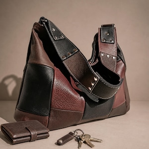 May include: A color-blocked hobo bag crafted from textured black, dark brown, and burgundy leather panels, featuring a wide shoulder strap secured with silver-toned metal rivets. A small matching brown leather wallet and a set of metal keys are included.