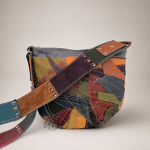 Puede incluir: Un bolso de hombro en forma de media luna hecho de parches de cuero multicolor en tonos verde oliva, amarillo mostaza, rojo oscuro, marrón y negro. El bolso presenta costuras blancas visibles y una correa de cuero ancha y multicolor.