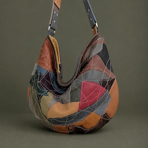 Pode incluir: Uma bolsa de ombro estilo hobo feita de um patchwork de peças de couro multicoloridas em tons de castanho, bege, vermelho, cinzento e verde. A bolsa apresenta costuras brancas visíveis num padrão curvo e uma alça de couro larga e ajustável.