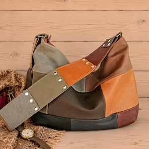 Può includere: Una borsa a spalla stile hobo realizzata con toppe in pelle multicolore nelle tonalità marrone, beige, verde oliva e nero. L'ampia tracolla presenta rivetti argentati e si collega al corpo principale con un design morbido e destrutturato.