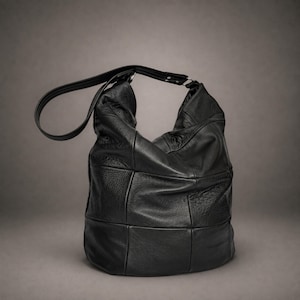 May include: A black hobo bag constructed from multiple panels of textured leather. The bag features a slouchy, rounded silhouette with a single wide shoulder strap attached by silver-toned metal hardware, showcasing a mix of smooth and pebbled finishes.