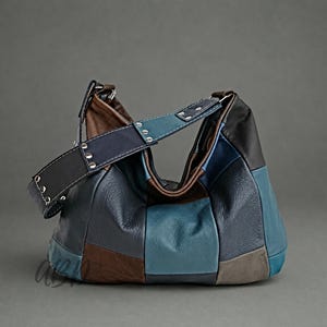 Può includere: Una borsa a spalla in stile hobo realizzata con toppe in pelle multicolore nei toni del blu navy, ottanio, marrone e grigio. La borsa presenta un'ampia tracolla regolabile fissata con rivetti in metallo argentato e una chiusura a zip.