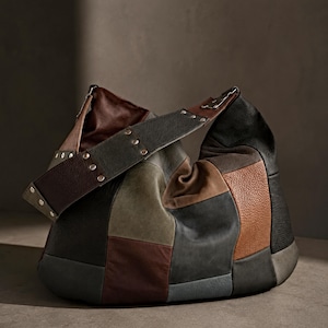 Könnte beinhalten: Eine Hobo-Tasche aus einem Patchwork aus Lederteilen in den Farben Schwarz, Braun, Olivgrün und Grau. Die Tasche verfügt über einen breiten, strukturierten Schulterriemen mit silberfarbenen Metallnieten und eine lockere, abgerundete Form.