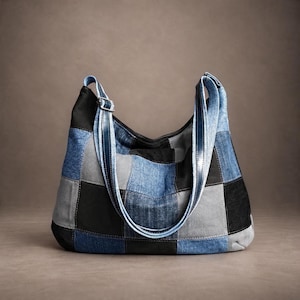 Puede incluir: Un bolso de hombro estilo hobo confeccionado con un mosaico de cuadrados de tela de mezclilla y pana en tonos negro, gris y azul. El bolso cuenta con dos correas de hombro largas y ajustables y un cierre de cremallera superior.