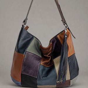 Könnte beinhalten: Eine Hobo-Schultertasche aus mehrfarbigen Lederflicken in Marineblau, Braun, Schwarz, Oliv und Beige. Die Tasche verfügt über einen langen, verstellbaren Riemen und eine lange, dunkle Lederquaste, die an einem silberfarbenen Metallverschluss befestigt ist.