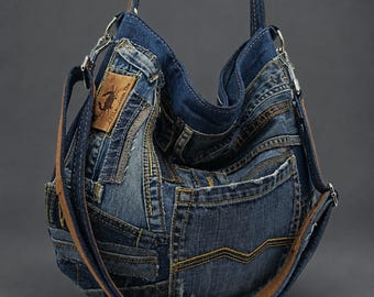 May include: A hobo-style shoulder bag constructed from repurposed blue denim fabric with visible seams, pockets, and frayed edges. The bag features a tan leather patch with a crab illustration and text, silver-tone metal hardware, and a brown leather strap.