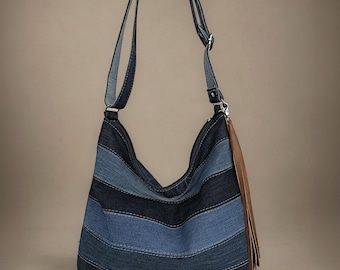 May include: A hobo-style shoulder bag constructed from horizontal panels of dark and medium blue denim fabric. The bag features a long adjustable strap and a prominent brown leather tassel attached to the side hardware with a silver metal clip.