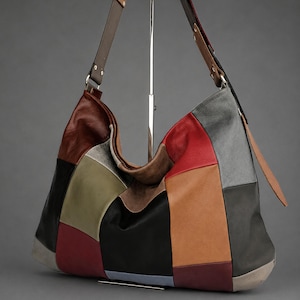 May include: A hobo-style shoulder bag constructed from multi-colored leather patches in shades of red, black, brown, gray, olive green, and tan. The bag features a wide, adjustable leather strap with metal hardware and a slouchy, unstructured shape.