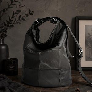 May include: Black leather shoulder bag with a patchwork design. The bag has a single shoulder strap and a top handle. The bag is sitting on a wooden surface. A black candle and a framed picture are in the background.