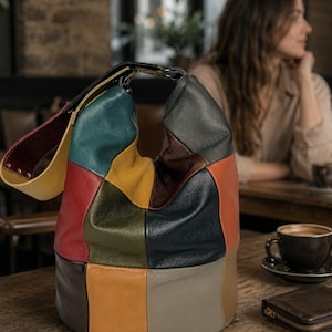 Op de afbeelding: Een veelkleurige patchwork leren handtas. De tas heeft panelen van rood, groen, geel en zwart leer. Het heeft een afgeronde vorm en een enkele schouderriem. De tas staat op een houten tafel.