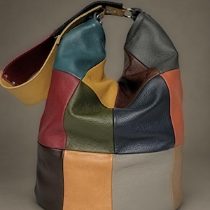 Puede incluir: Un bolso tipo hobo confeccionado con paneles de cuero texturizado multicolor en tonos azul verdoso, amarillo, verde oliva, marrón oscuro, azul marino, naranja, gris y beige. El bolso cuenta con una correa ancha ajustable con herrajes plateados.