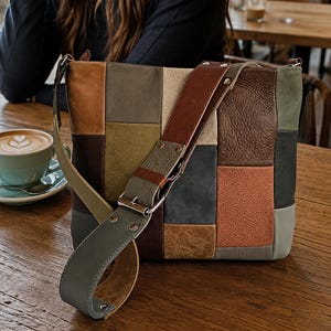 Könnte beinhalten: Eine rechteckige Umhängetasche aus einem Patchwork aus Lederstücken in verschiedenen Farben, darunter Braun, Beige, Olivgrün, Grau und Terrakotta. Die Tasche verfügt über einen breiten, verstellbaren Lederriemen mit silberfarbenen Metallbeschlägen.