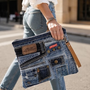 May include: A rectangular clutch bag constructed from various shades of blue denim fabric patches. The bag features a brown leather patch with the text Jerem UPPER CASUAL JEANS, a small red label, metal buttons, and a tan leather wrist strap.