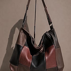 May include: A hobo-style shoulder bag crafted from panels of pebbled leather in a patchwork design. The bag features alternating sections of black, dark brown, and deep burgundy leather, with a single wide shoulder strap and a slouchy silhouette.