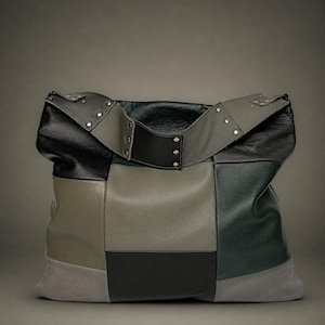 Op de afbeelding: Een grote, slappe tote bag gemaakt van een patchwork van leren panelen in tinten zwart, antracietgrijs en olijfgroen. De brede schouderband is voorzien van zilverkleurige metalen klinknagels en de tas heeft een gestructureerd ontwerp met meerdere panelen.
