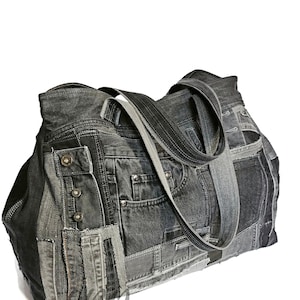 May include: A large, patchwork denim tote bag in shades of grey. The bag features multiple denim panels, pockets, and button details. It has two long straps for carrying over the shoulder or by hand. The bag is set against a white background.
