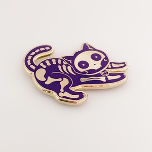 May include: Gold-coloured enamel pin of a purple cat skeleton with a pentagram charm. The cat is in a playful pose with its tail curled around its body.