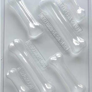 May include: Clear plastic bone-shaped candy mold. The mold has six bone shapes of varying sizes. The text "CONCEPTS IN CANDY", "NORTHRIDGE, CA 91324", "FOOD APPL MAT", and "B127©2006" are visible.