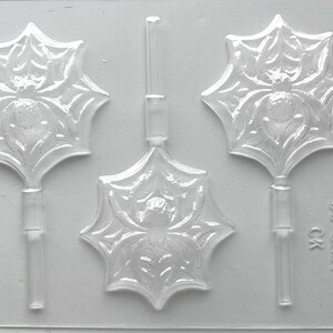 May include: A clear plastic mold for making spider-shaped lollipops. The mold features three spider designs with intricate web details. The mold is designed for easy candy making and is ideal for Halloween treats.