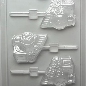 May include: A clear plastic mold for making train-shaped lollipops or chocolates. The mold has three cavities, each with a detailed train design, including wheels, a smokestack, and other features. The mold is designed to hold the lollipop sticks.