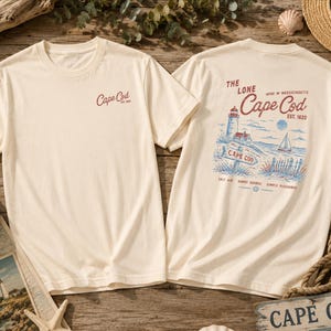 May include: A cream-colored short-sleeve t-shirt featuring a small red Cape Cod logo on the front left chest. The back displays a detailed graphic of a lighthouse, sailboat, and coastal scenery with the text The Lone Cape Cod Est. 1620.
