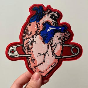 May include: Embroidered patch featuring a stylized anatomical heart in shades of pink, blue, and white, pierced by a red safety pin. The heart has detailed line work and is outlined in red.