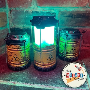 May include: Three yellow and black metal lanterns with a radioactive symbol on the side. The lanterns are lit with a green glow. The lanterns are on a brick surface. The text "The Mojave Outpost" is on a round sticker with a rocket ship.