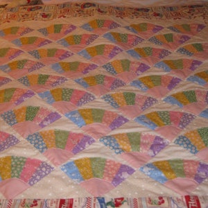 May include: A white quilt with a pattern of pink, purple, blue, and green fan-shaped pieces. The quilt is made of patchwork fabric and has a white background with small white dots.