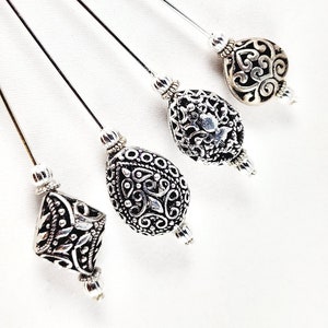 May include: Four silver hair pins with decorative, ornate, filigree bead tops. The beads are different shapes and sizes.