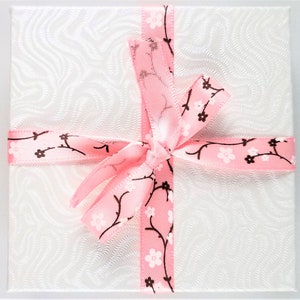 May include: A white gift box with a pink ribbon tied in a bow. The ribbon has a pattern of brown branches with pink flowers.