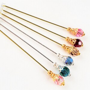 May include: Six gold-toned hat pins with decorative tops and various colored crystal beads. The beads are pink, red, brown, blue, teal, and clear.