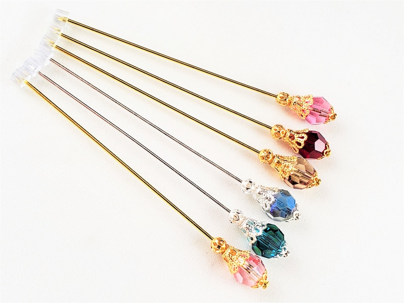 May include: Six gold-toned hat pins with decorative tops and various colored crystal beads. The beads are pink, red, brown, blue, teal, and clear.