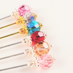 May include: Six decorative pins with colorful crystal beads and gold or silver accents. The beads are pink, clear, teal, red, amber, and pink.