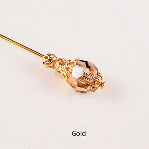 May include: A gold-toned hat pin with a clear glass teardrop-shaped bead and a decorative metal design. The word "Gold" is written below the pin.