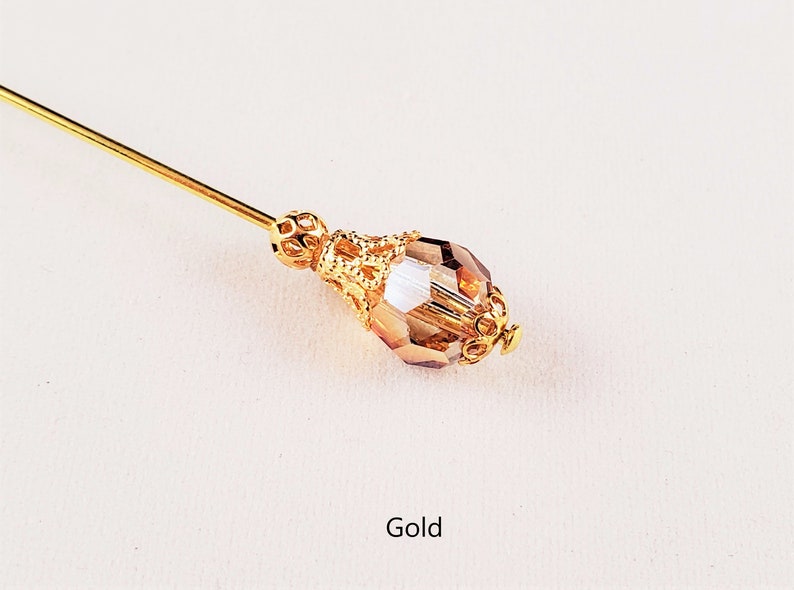 May include: A gold-toned hat pin with a clear glass teardrop-shaped bead and a decorative metal design. The word "Gold" is written below the pin.