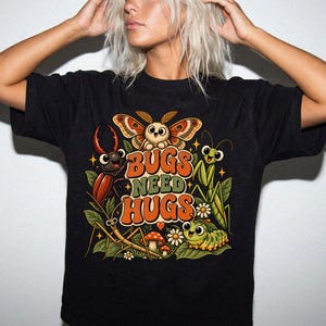 May include: A black t-shirt featuring a colorful graphic of various cartoon insects, including a moth, beetle, mantis, and caterpillar, surrounded by leaves, flowers, and mushrooms. The text "BUGS NEED HUGS" is printed in large, bold, orange letters.