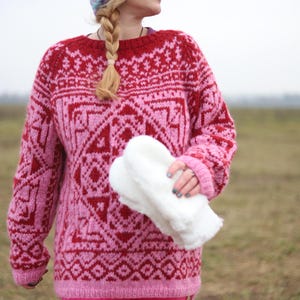 May include: A woman wearing a pink and red patterned sweater with a white pom-pom hat. She is holding a white fluffy item in her hands.