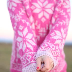Könnte beinhalten: Ein leuchtend pinker Pullover mit Stehkragen und einem sich wiederholenden weißen Schneeflocken- und geometrischen Muster auf Brust und Ärmeln. Das Strickkleidungsstück verfügt über gerippte Bündchen und einen gerippten Halsausschnitt.