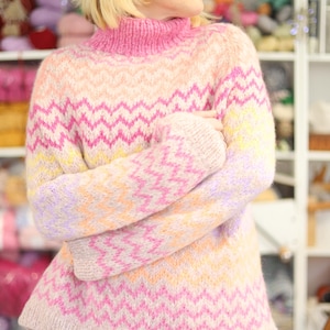 May include: A colorful, hand-knitted sweater with a high neck and a zig-zag pattern in shades of pink, peach, yellow, and lavender. The sweater is made of soft, fluffy yarn and has long sleeves. The model is wearing white pants.