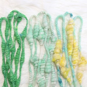 Puede incluir: Primer plano de varios hilos de lana colorida y texturizada. La lana es en tonos de verde, azul y amarillo, con un aspecto suave y difuso. Los hilos están dispuestos sobre un fondo blanco y esponjoso, creando una exhibición visualmente atractiva.