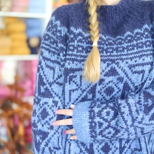 May include: A blue sweater with a geometric pattern in a lighter shade of blue. The sweater has a high neckline and long sleeves. The design is a repeating pattern of diamonds and other shapes. The sweater is a cozy, handmade item.