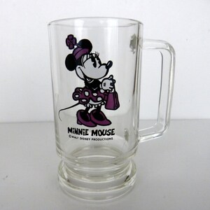 Może przedstawiać: Przezroczysty szklany kubek z uchwytem, z Minnie Mouse w fioletowej sukience i kokardzie. Kubek ma napisy "Minnie Mouse" i "Walt Disney Productions".