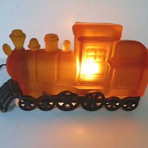 May include: A frosted orange glass train lamp with a warm internal glow. The locomotive features a detailed cab, chimney, and boiler, mounted on a dark metal base with six spoked wheels and a front cowcatcher. A black power cord extends from the side.