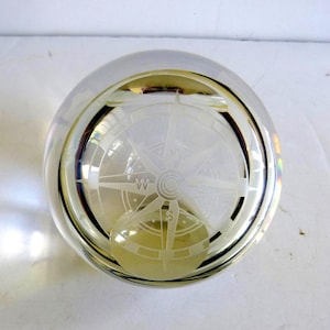 May include: A clear, spherical glass paperweight featuring an etched compass rose design on its surface. The compass rose includes cardinal direction markers, with the letter W clearly visible on the left side and S on the bottom section.