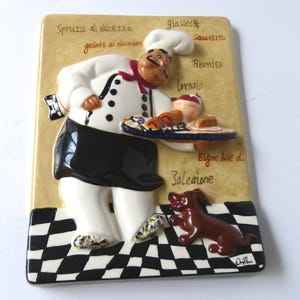 May include: A ceramic wall plaque featuring a chef holding a tray of food, with a dog at his feet. The chef is wearing a white uniform and black apron. The plaque has Italian text and a black and white checkered base.