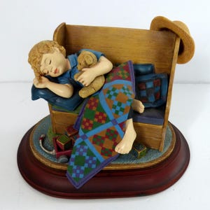 May include: A figurine depicting a sleeping child holding a teddy bear, resting on a wooden bench. The child is covered with a colorful quilt. A straw hat rests on the bench's back. The figurine is set on a round, dark red base.