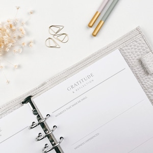 May include: A light grey leather planner, open to a page with the words "Gratitude & Reflection". The page has lines for writing. Gold paper clips and pens with gold tips are also visible. The planner is on a white surface.