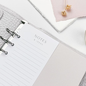 May include: A light grey binder with silver rings, holding lined paper with the text "NOTES & IDEAS." A roll of decorative tape with a floral design and dried flowers are also visible. The image suggests a stationery and organisation theme.